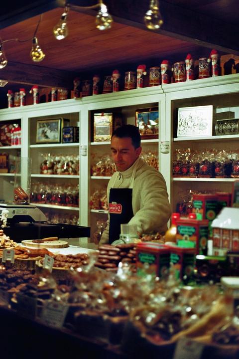 A man wearing an apron stands behind a counter filled with various baked goods and candies, with shelves of packaged items and Santa figurines in the background.