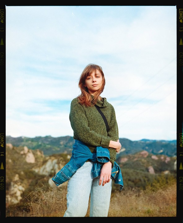 A woman wearing a green sweater and a denim jacket tied around her waist stands in a hilly, dry landscape under a bright sky.