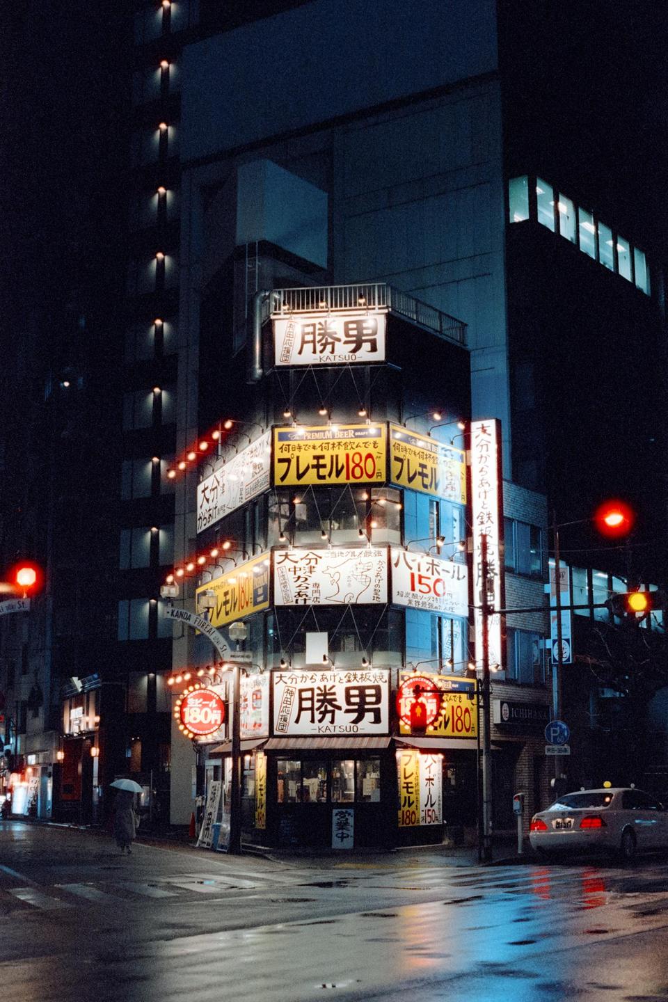 A street corner building with many illuminated signs is seen at night with a person holding an umbrella and a car on the wet street.