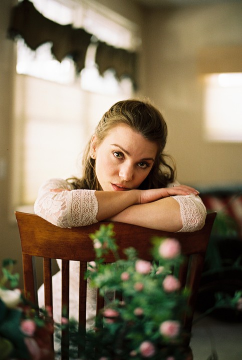 A woman in a lace top leans on the back of a wooden chair, looking directly at the viewer with blurred pink flowers in the foreground.