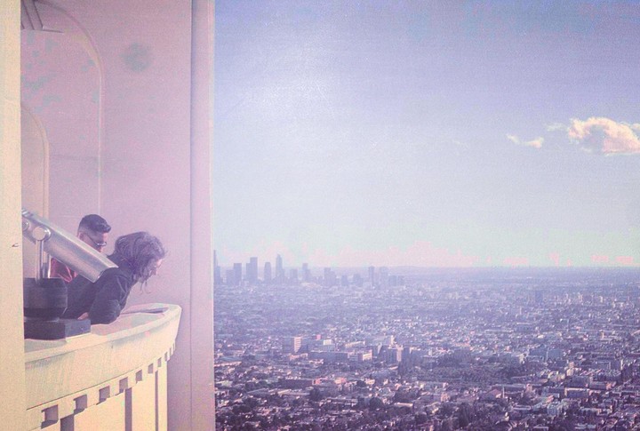 A woman and a man look out from a balcony over a hazy city skyline.