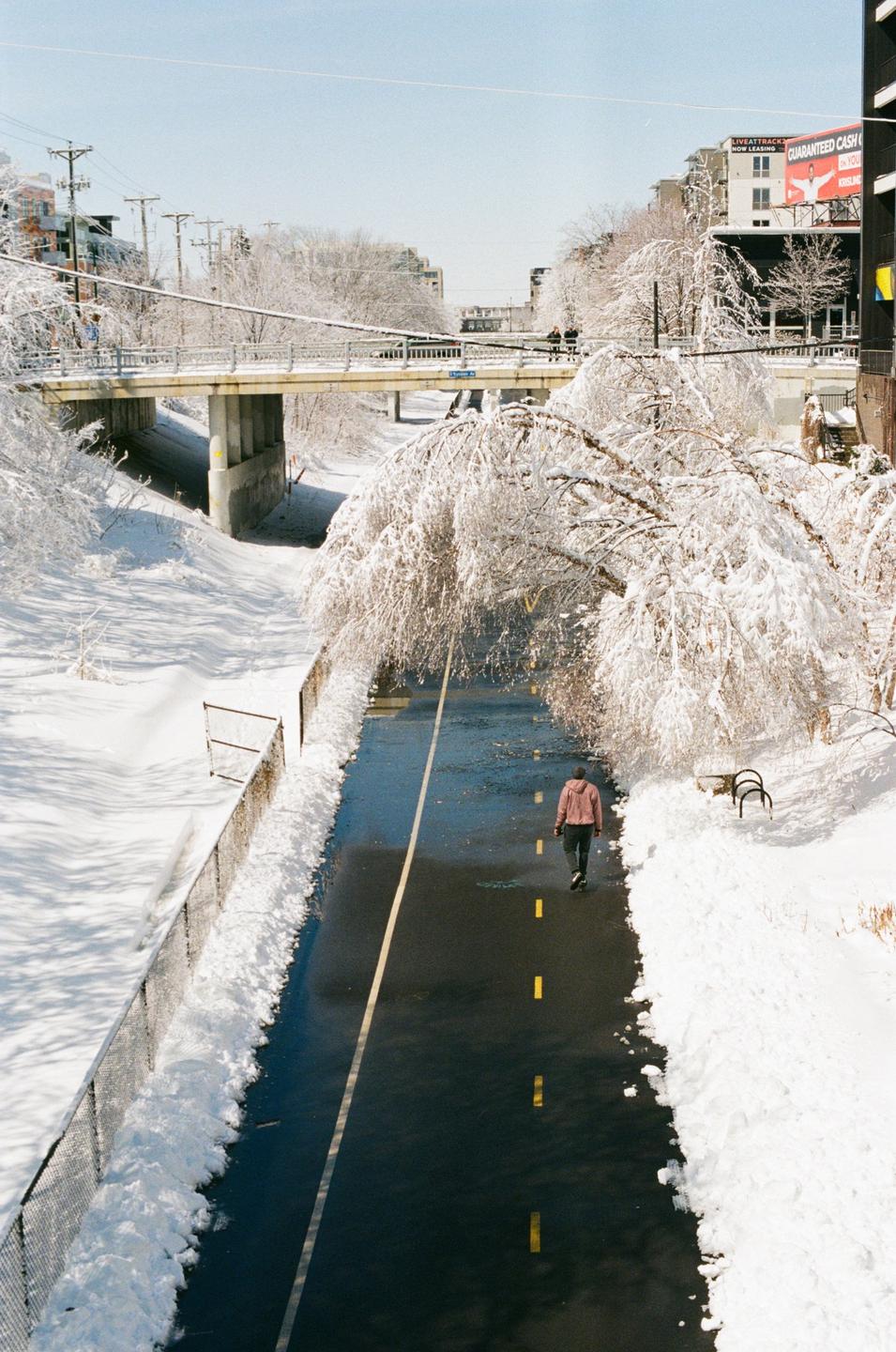A person walks along a path cleared of snow, with snow-covered trees and a bridge overhead.