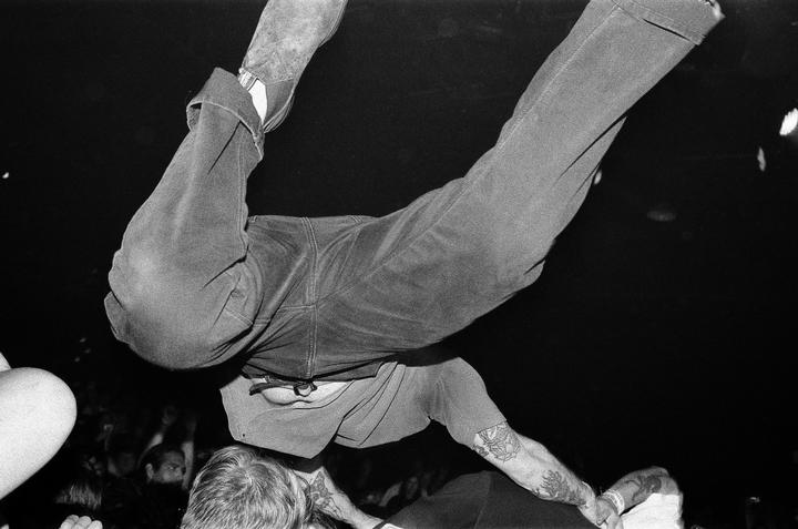 A man in jeans and a t-shirt is crowd surfing at a punk show, with his legs and torso visible from an unusual angle.