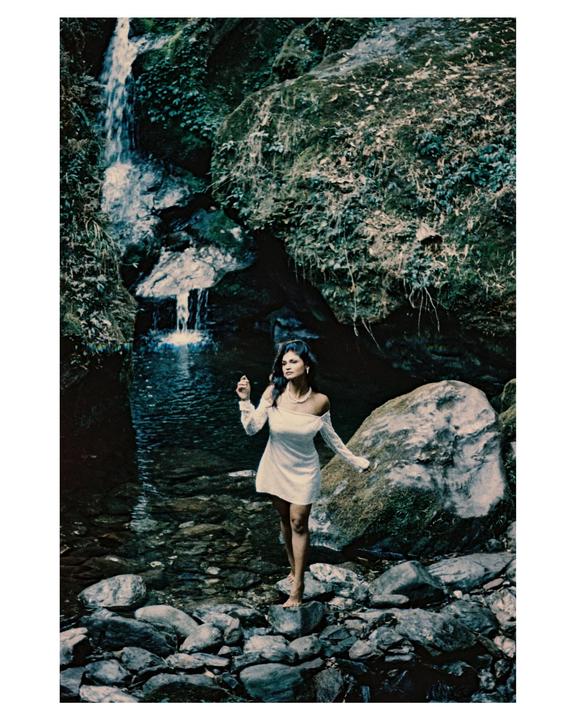 A woman in a white dress stands barefoot on rocks in a stream with a waterfall in the background.