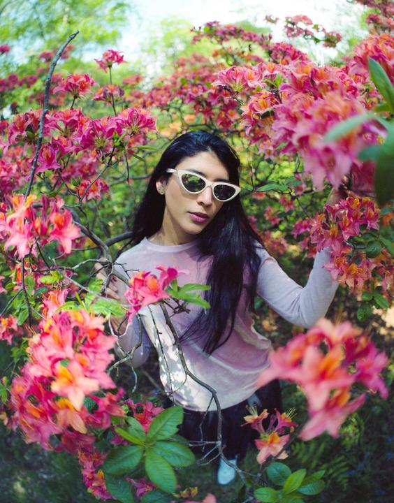 A woman wearing sunglasses stands among bright pink and orange flowers.