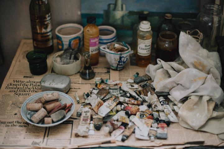 A collection of art supplies, including paint tubes, bottles, and brushes, are arranged on a newspaper-covered surface.
