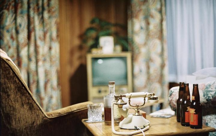 A rotary telephone, a glass, a bottle of liquor, and three beer bottles sit on a wooden table next to an armchair, with a television and patterned curtains in the background.