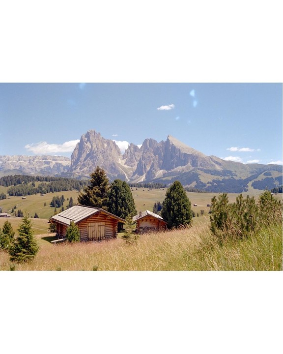 Two wooden cabins sit in a field of tall grass with a mountain range in the background under a blue sky.