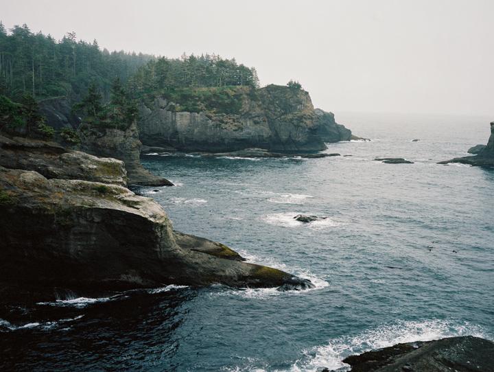 A rugged coastline with cliffs and evergreen trees meets the ocean under a hazy sky.