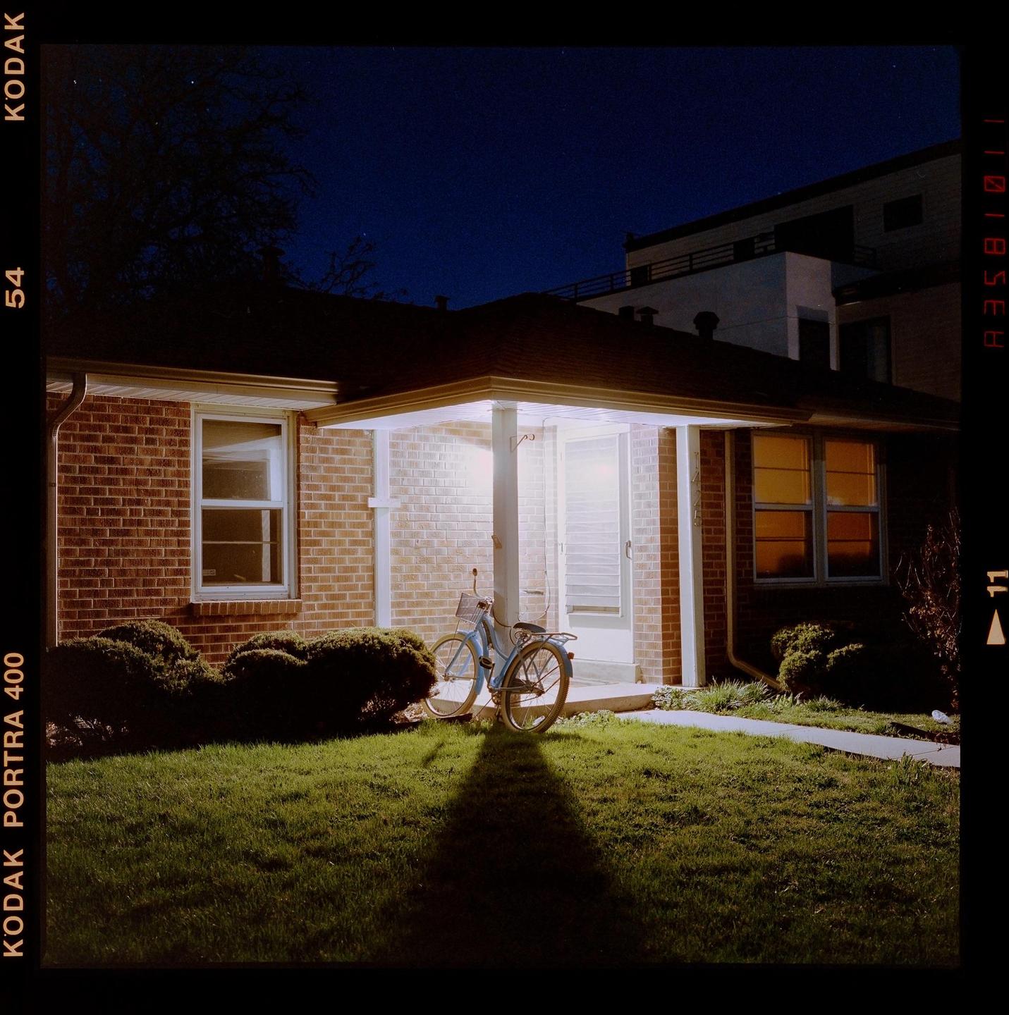 A bicycle is parked on a lawn in front of a brick house with a lit porch at night.