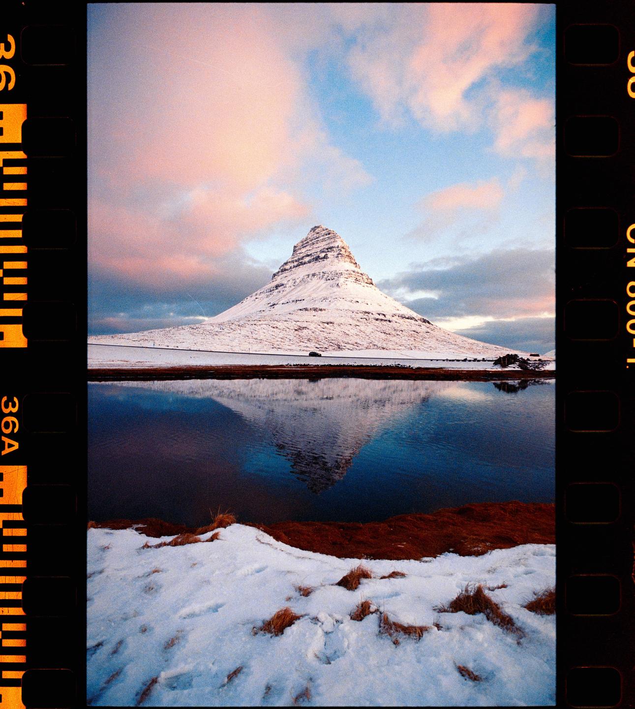 A snow-covered mountain reflects in a body of water under a sky with pink clouds.
