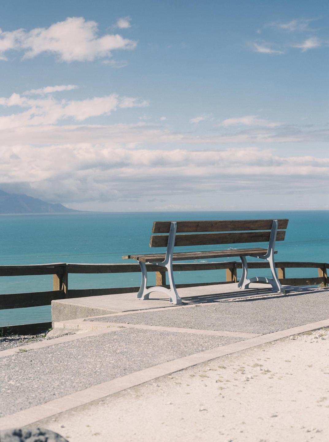 A wooden bench with a metal frame sits on a paved area overlooking a large body of water with mountains in the distance under a cloudy sky.