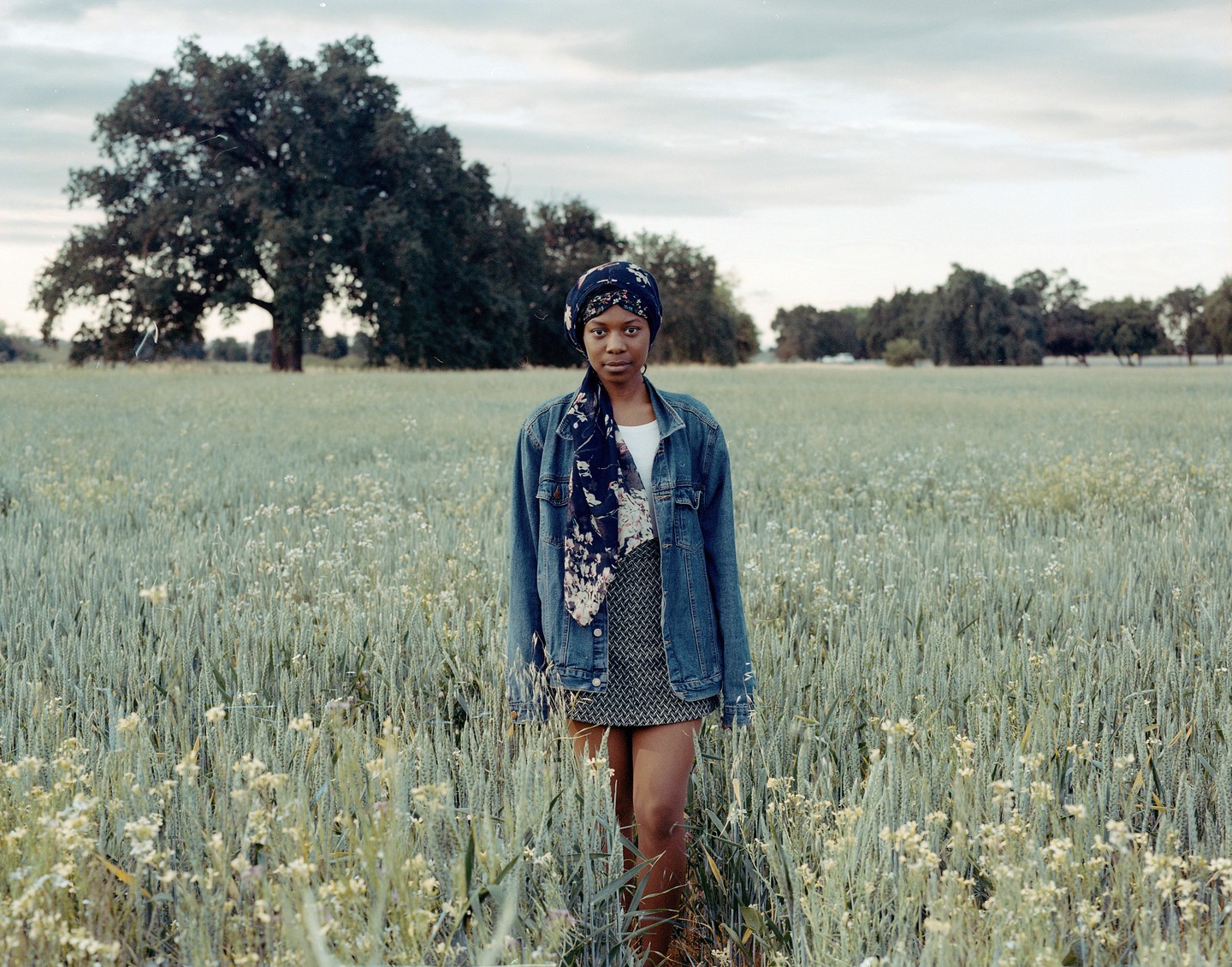 A woman stands in a field of tall grass and wildflowers with trees in the background under a cloudy sky.