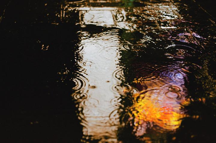 Raindrops create ripples on a wet, reflective surface at night, with colorful light reflections.