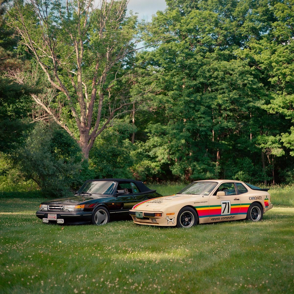 Two cars are parked on a grassy field in front of a forest.
