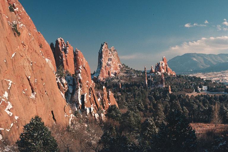 Red rock formations covered in snow rise above a forest with mountains in the distance under a blue sky.