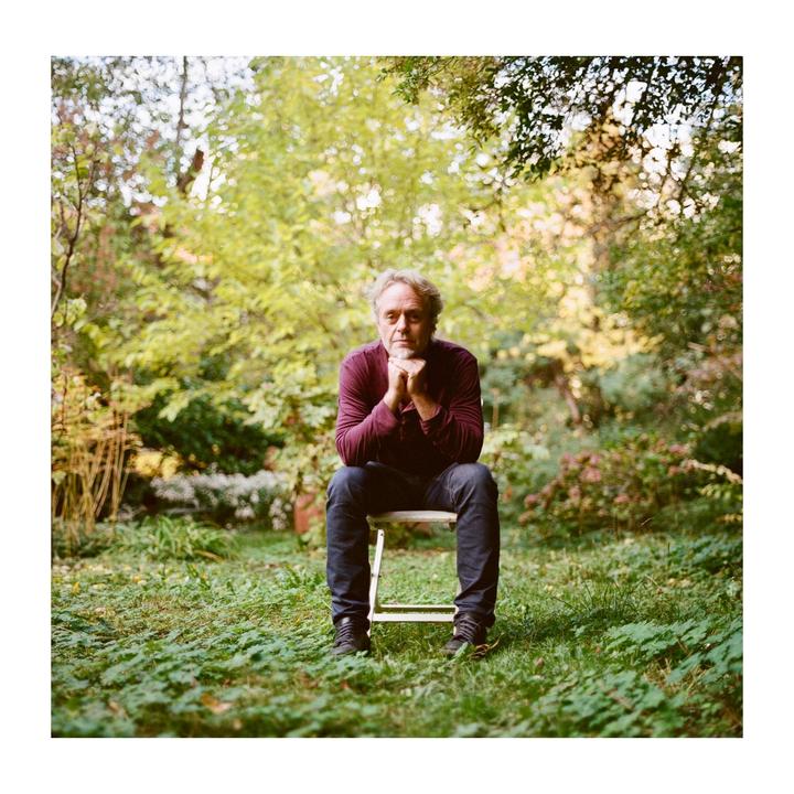 A man with gray hair and a beard sits on a chair in a garden with his hands clasped under his chin.