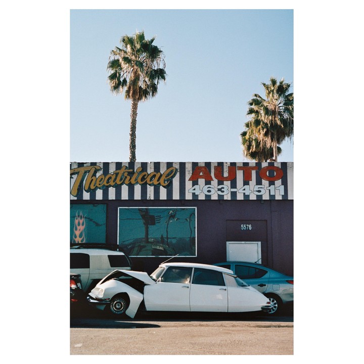 A white car with a crumpled hood is parked in front of a building with a sign that reads "Theatrical Auto" and two palm trees.