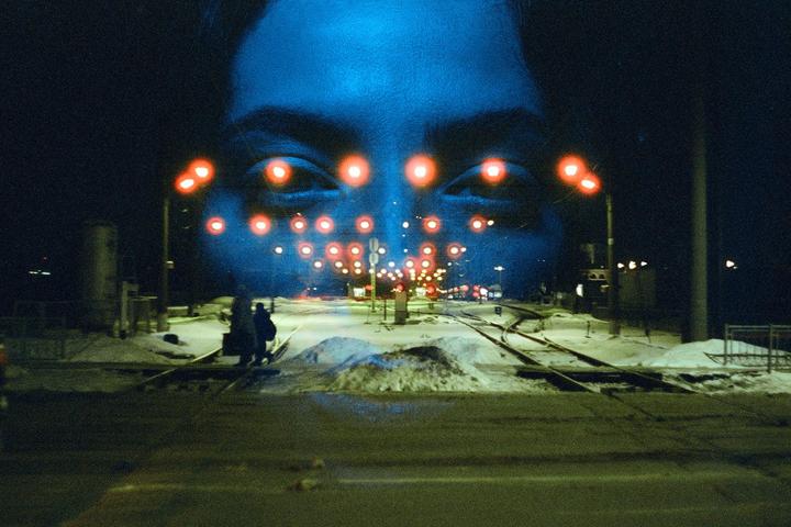 A large blue face with red lights for eyes is double exposed over a snowy street with railroad tracks at night.