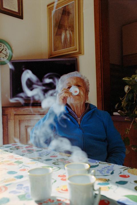 A woman sips from a small cup at a table with a patterned tablecloth, with steam rising from other cups in the foreground.