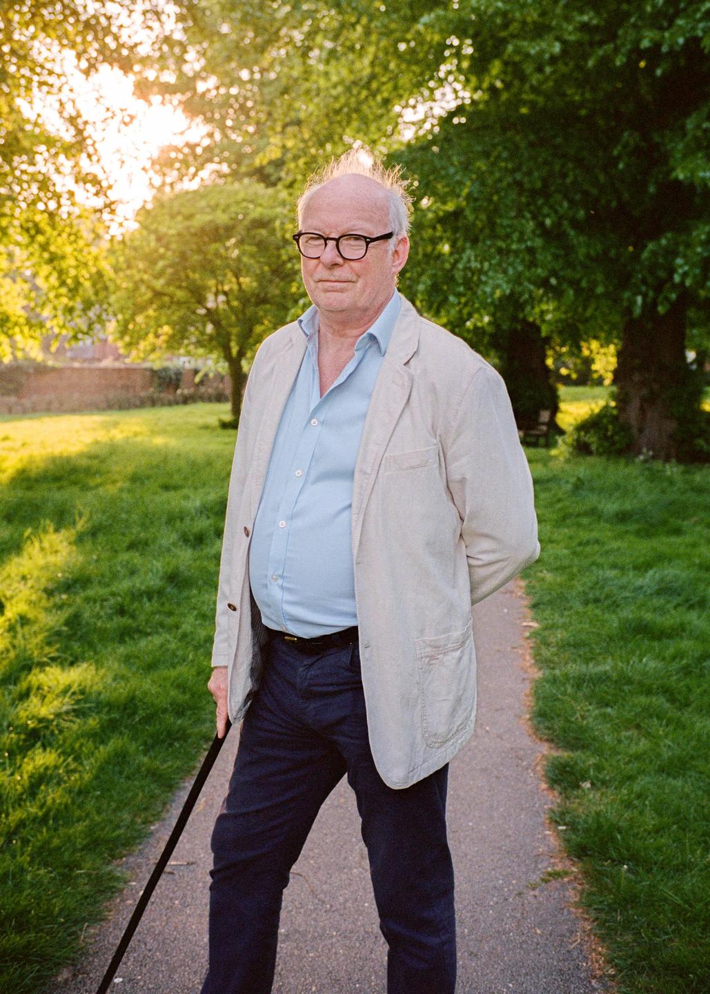 A man wearing glasses, a light blue shirt, and a light jacket stands on a path in a park with trees and grass, holding a cane.