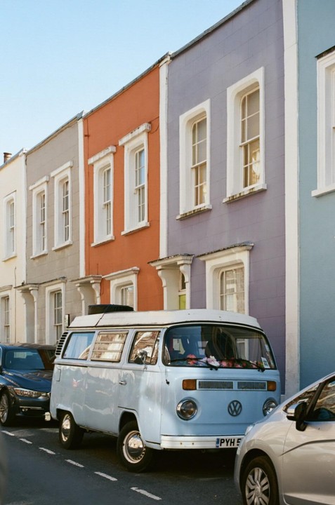 A light blue vintage van is parked on a street lined with colorful terraced houses.