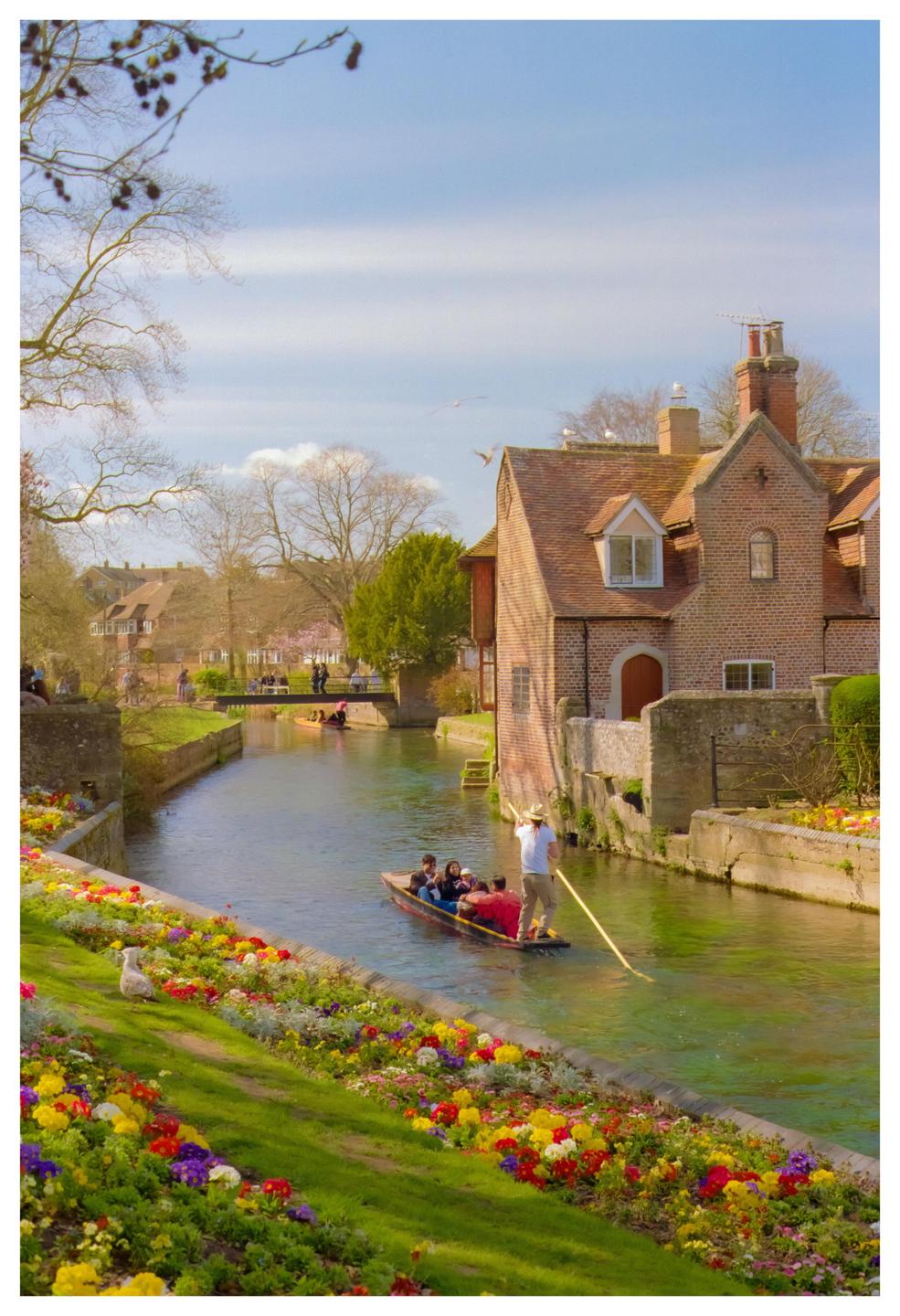 A man punts a boat with people down a river lined with colorful flowers and historic buildings.