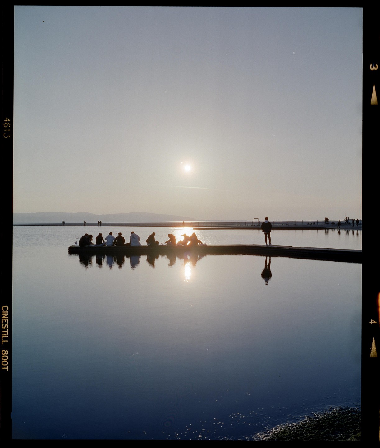 A group of people sit on a pier watching the sunset over a calm body of water, with one man standing alone further down the pier.