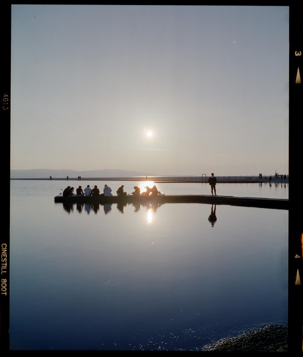 A group of people sit on a pier watching the sunset over a calm body of water, with one man standing alone further down the pier.