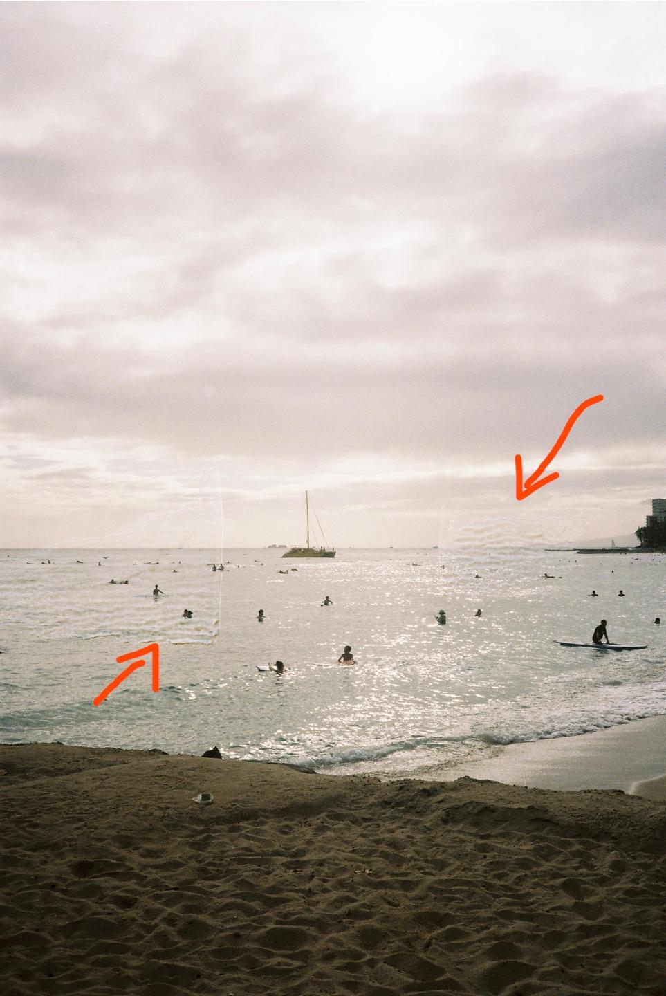 A beach scene shows many people swimming and surfing in the ocean with a boat in the distance under a cloudy sky.