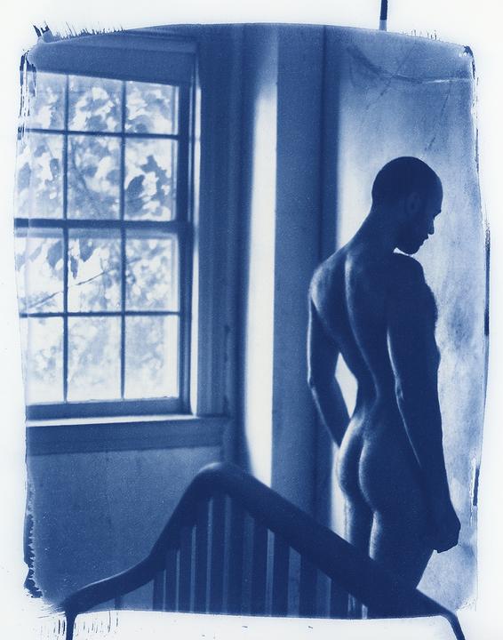 A nude man stands next to a window with a bed frame in the foreground.