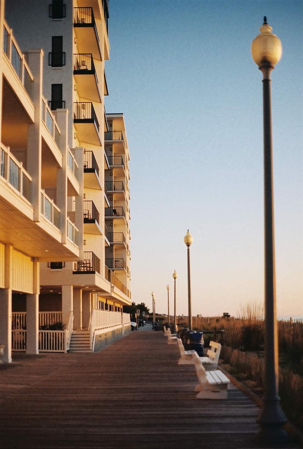 A boardwalk with benches and streetlights runs alongside a tall building with balconies, with a beach and ocean visible in the distance.