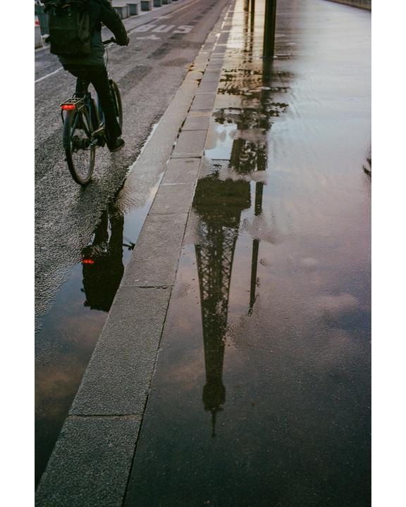 A person rides a bicycle on a wet street next to a puddle reflecting the Eiffel Tower.