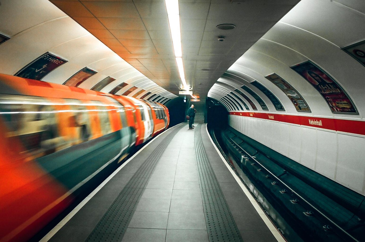 A man stands on a subway platform as a train speeds past, creating a motion blur effect.