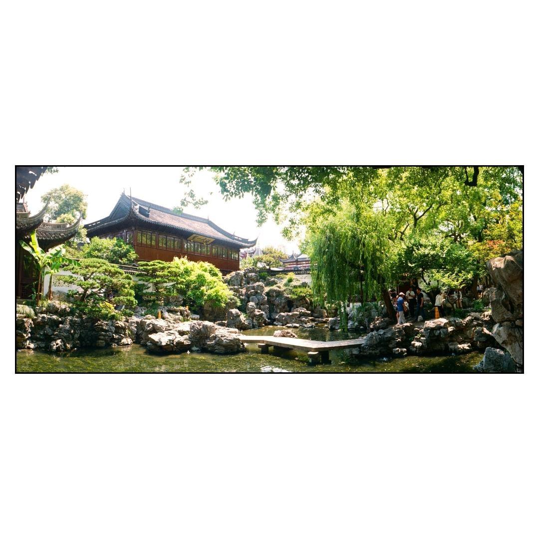 A traditional Chinese building with a dark roof stands behind a pond with rocks and lush green trees.