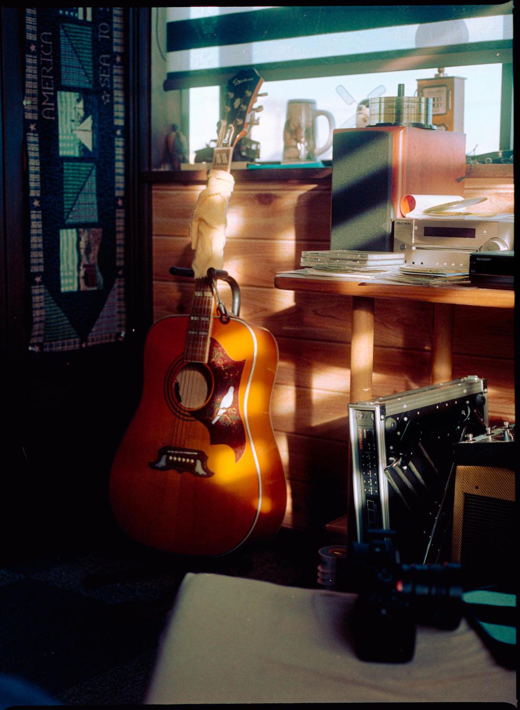 An acoustic guitar stands in a sunlit room next to a wooden table with various electronics.
