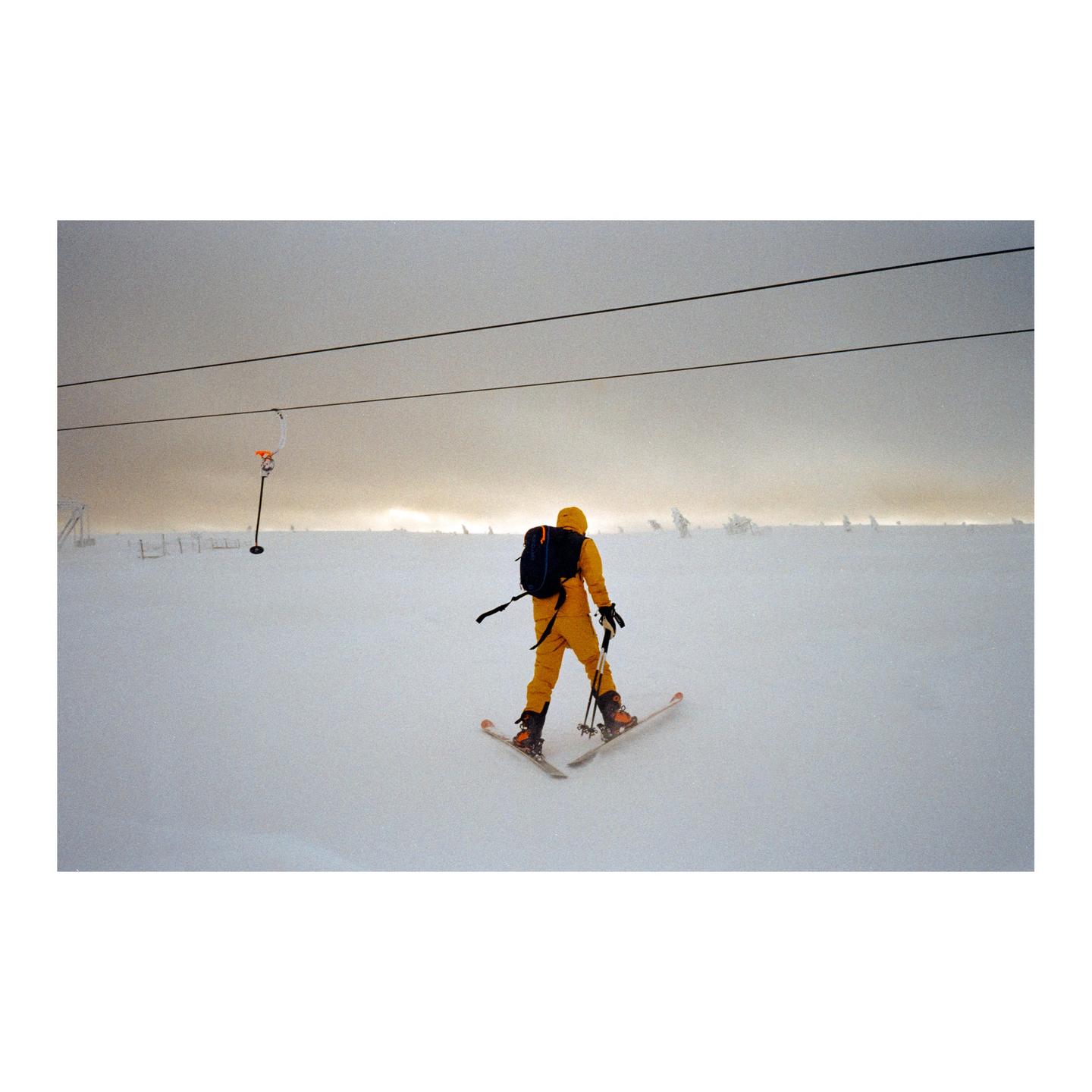 A person in a yellow ski suit skis down a snowy slope under a cloudy sky.