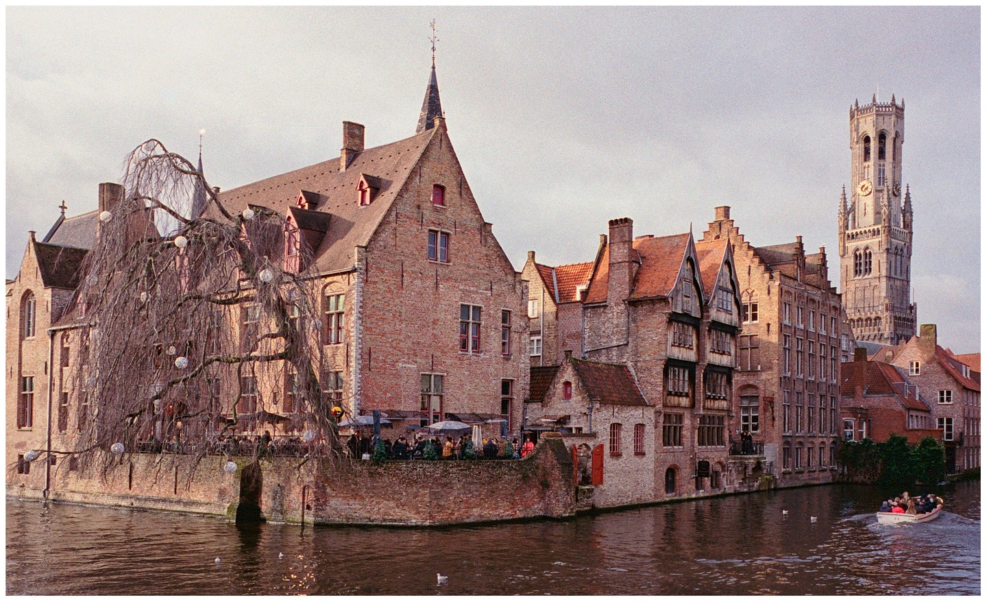 A canal with a boat passes by historic brick buildings and a tall bell tower under a cloudy sky.