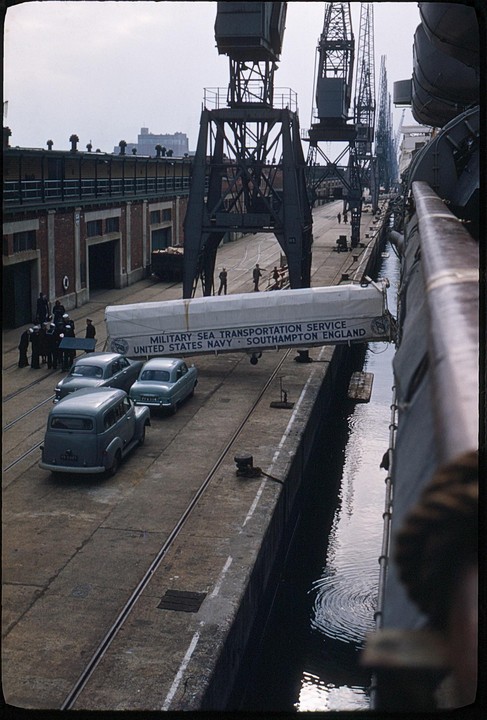 A gangway with text about military sea transportation connects a ship to a dock where cars are parked and cranes are visible.