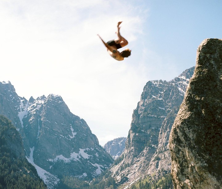 A man performs a backflip in the air with mountains in the background.