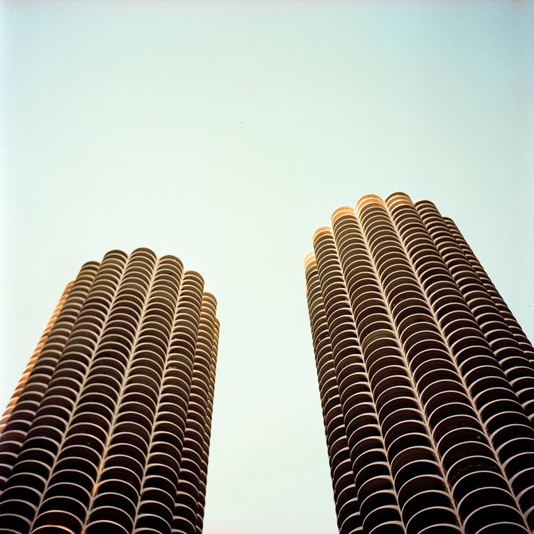 Two tall, cylindrical buildings with many balconies are viewed from below against a clear sky.