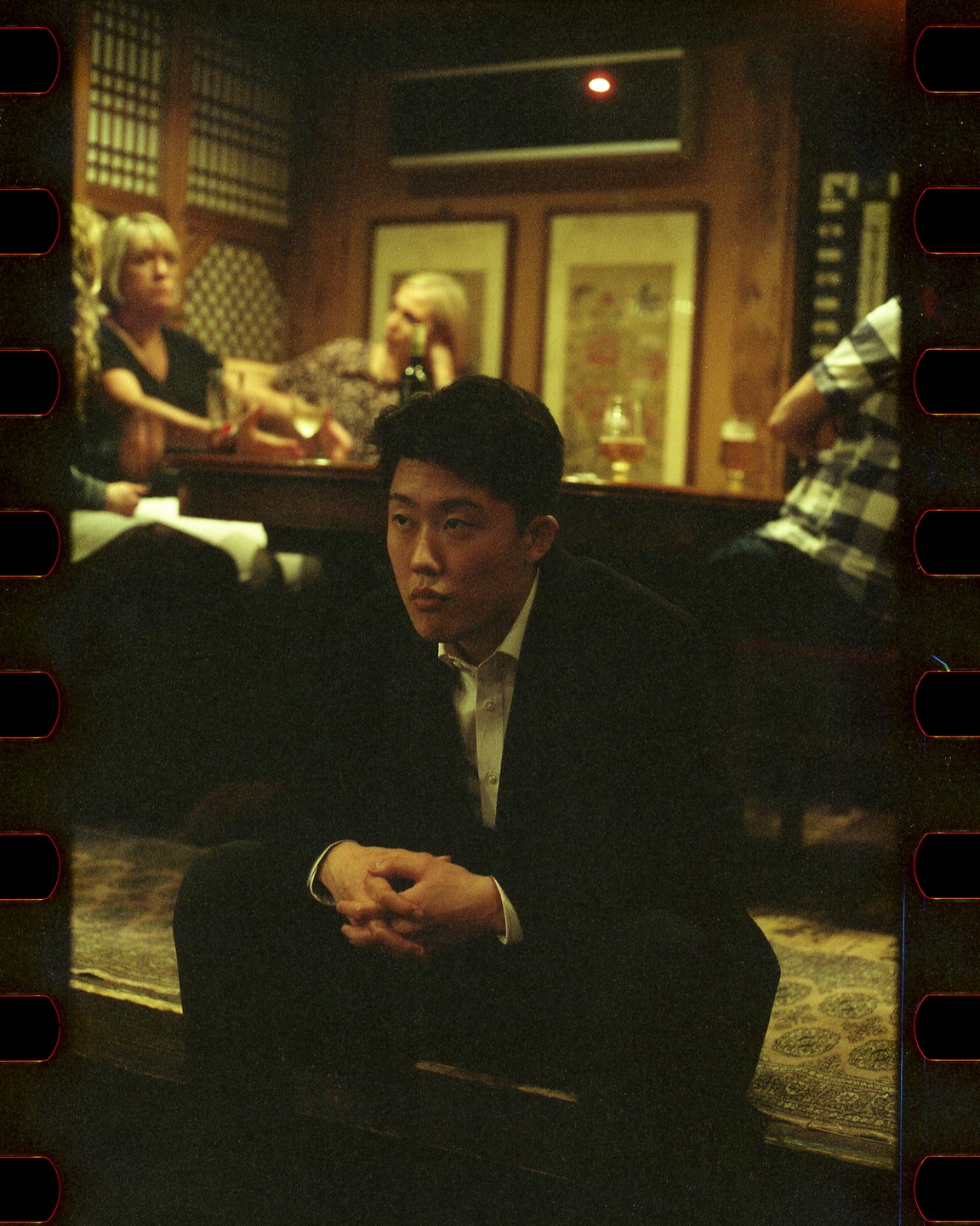 A man in a suit sits on a step with his hands clasped, looking to the side, with other people and drinks visible in the background.