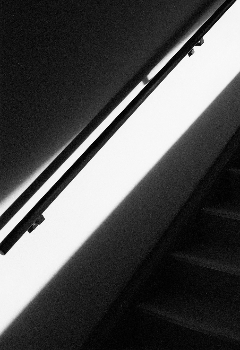 A black handrail casts a long shadow on a white wall next to a dark staircase.