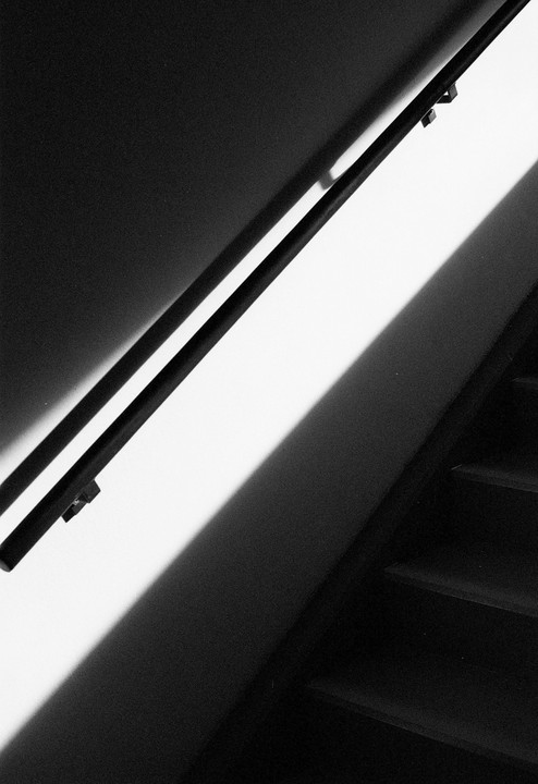 A black handrail casts a long shadow on a white wall next to a dark staircase.