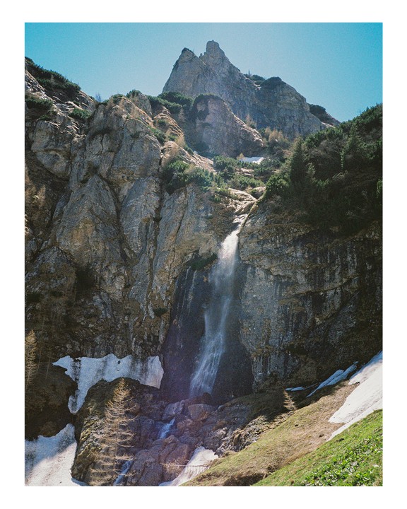 A waterfall cascades down a rocky mountain with patches of snow and green grass.