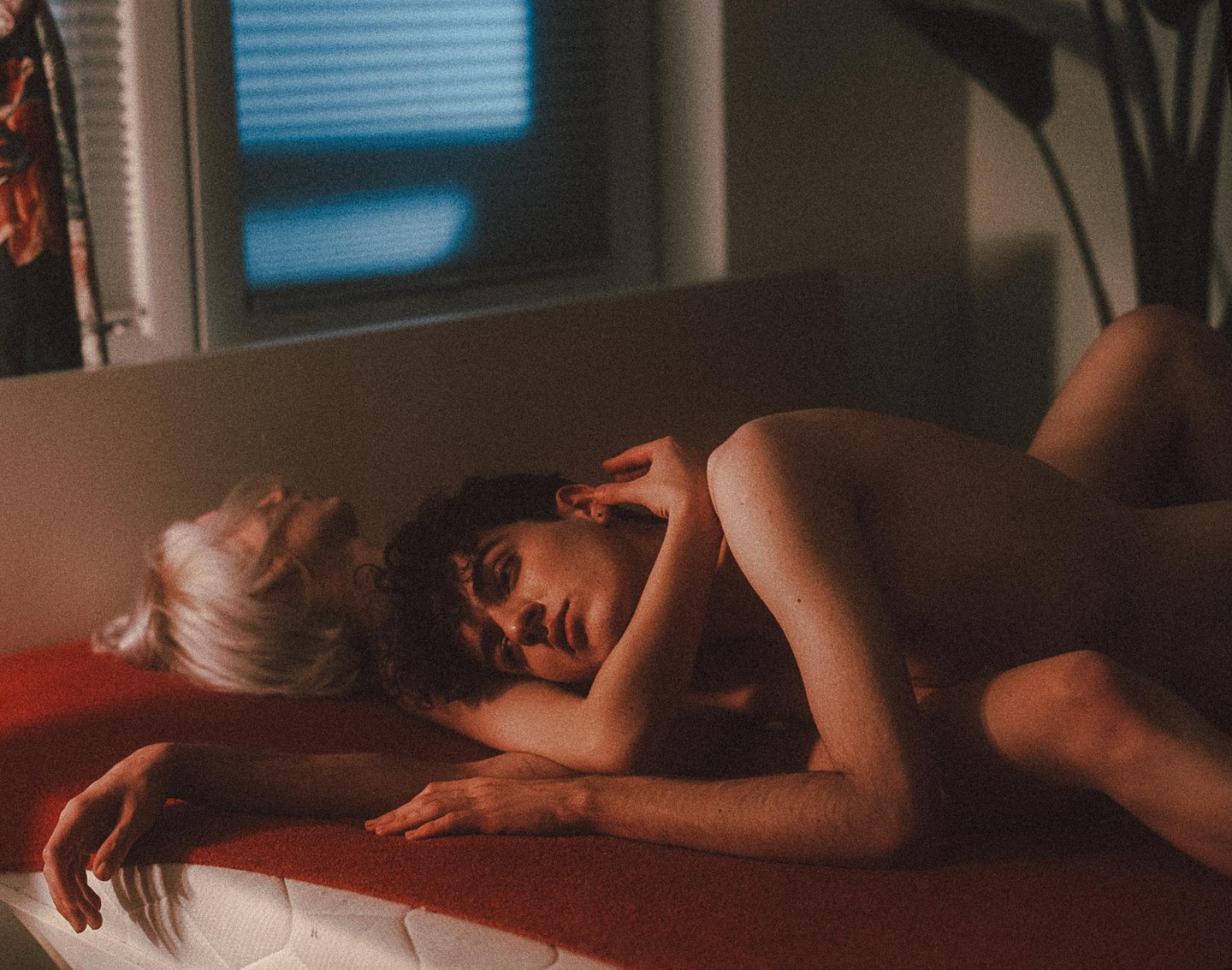 A couple lies together on a red blanket, with one person looking towards the viewer and the other looking away.