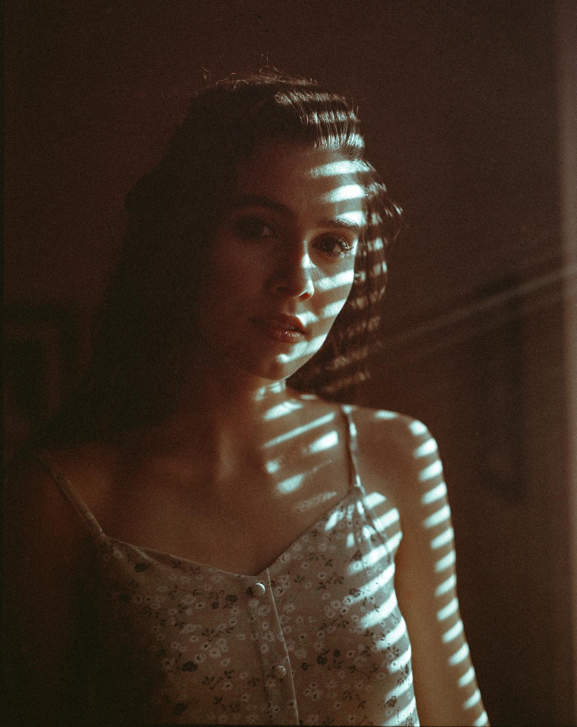 A woman with dark hair looks at the viewer, with shadows from blinds falling across her face and shoulder.