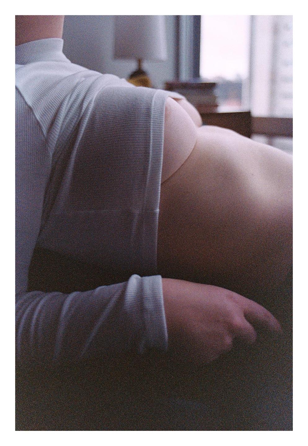 A woman is seen from the back, partially clothed in a light-colored top, with a lamp and window in the background.