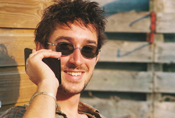 A man wearing sunglasses and a plaid shirt smiles while holding a phone to his ear.
