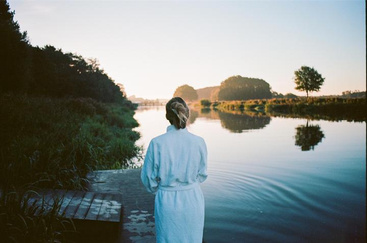 A woman in a bathrobe stands on a wooden dock looking out at a river at dawn.